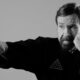 Homem idoso, Chuck Norris, com barba, soca saco de pancadas. Foto em preto e branco.