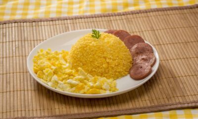 Prato branco com cuscuz de milho, ovos mexidos e linguiça fatiada sobre esteira de bambu.