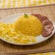 Prato branco com cuscuz de milho, ovos mexidos e linguiça fatiada sobre esteira de bambu.