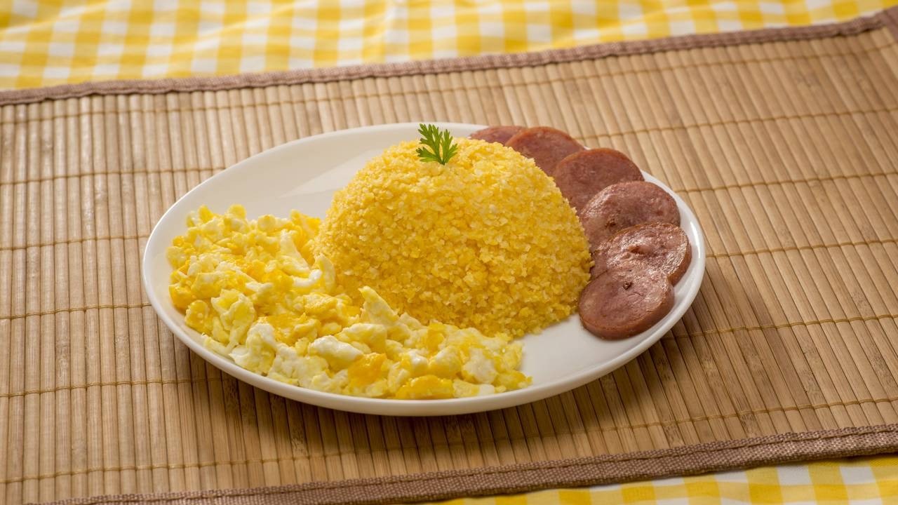 Prato branco com cuscuz de milho, ovos mexidos e linguiça fatiada sobre esteira de bambu.