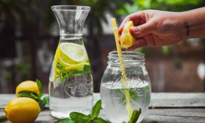 Mão espremendo limão em copo com água, canudo; jarra e limões frescos na mesa.