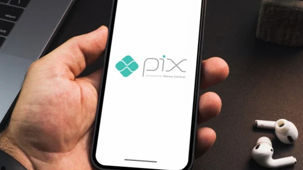Mão segura smartphone com o logo do Pix na tela, ao lado de fones de ouvido brancos.