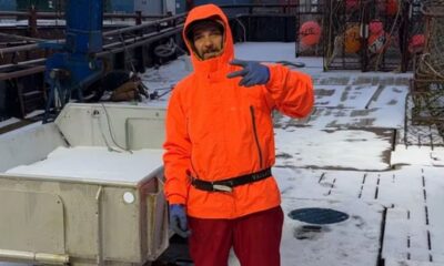 Jovem de jaqueta laranja e luvas azuis faz sinal de paz em barco coberto de neve.