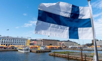 Bandeira da Finlândia tremulando sobre porto com barcos, tendas e edifícios à beira-mar.