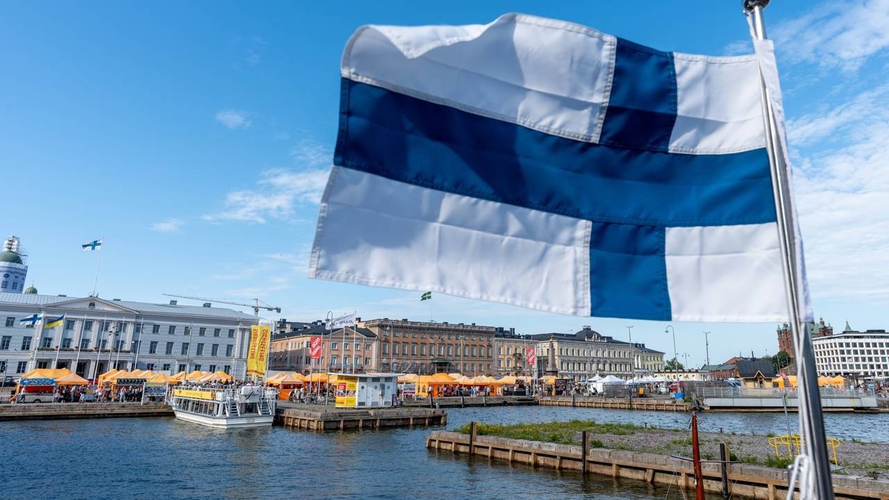 Bandeira da Finlândia tremulando sobre porto com barcos, tendas e edifícios à beira-mar.