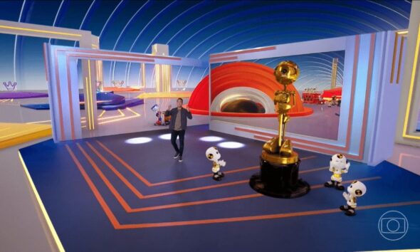 Tadeu Schmidt gesticula em estúdio azul com troféu dourado, quatro robôs e tela com cenário laranja.