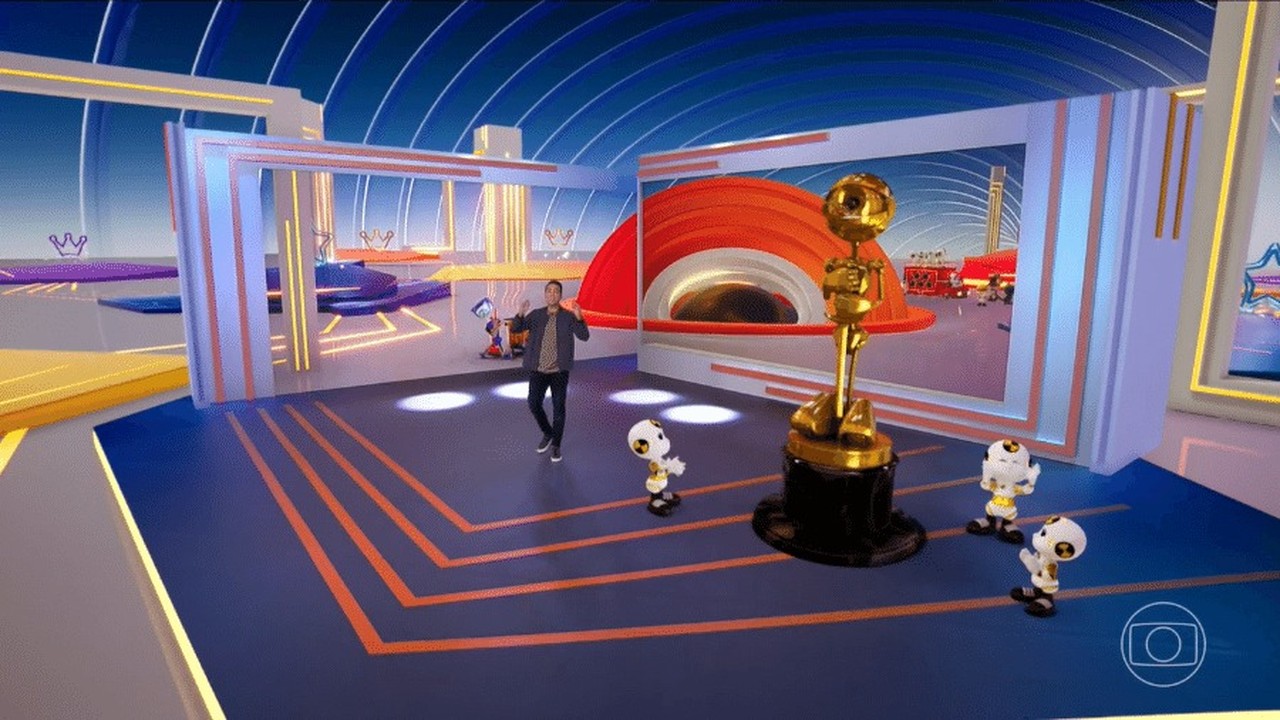 Tadeu Schmidt gesticula em estúdio azul com troféu dourado, quatro robôs e tela com cenário laranja.