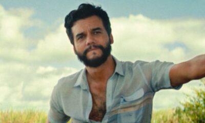 Retrato de Wagner Moura, homem barbudo, camisa azul aberta, olhando. Fundo de campo e céu.