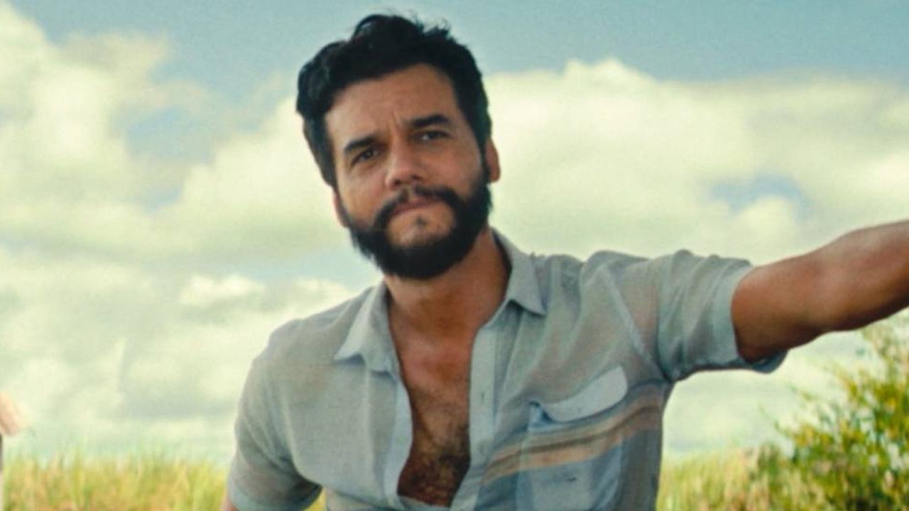 Retrato de Wagner Moura, homem barbudo, camisa azul aberta, olhando. Fundo de campo e céu.