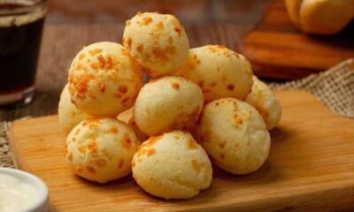 Pão de queijo recém-assado empilhado em tábua de madeira com café e molho.