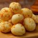 Pão de queijo recém-assado empilhado em tábua de madeira com café e molho.