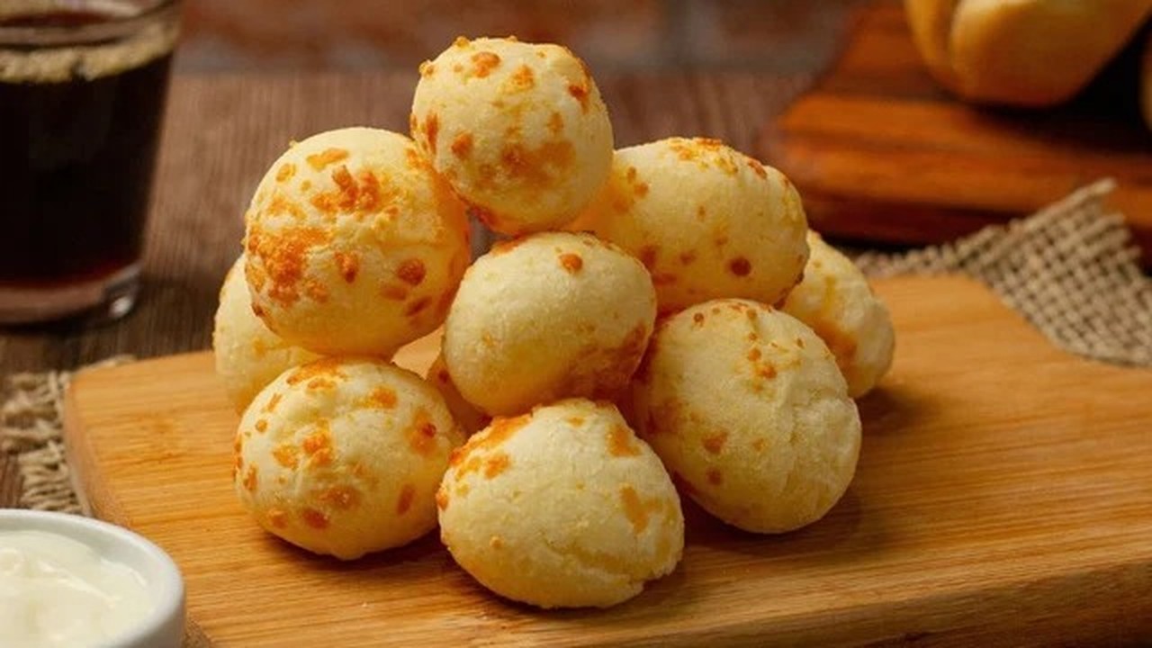 Pão de queijo recém-assado empilhado em tábua de madeira com café e molho.