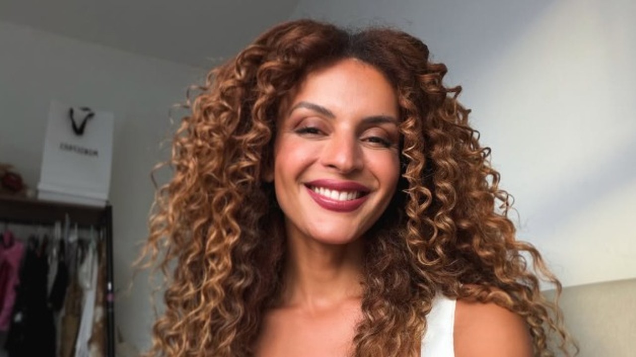 Gabriela Loran sorri para a câmera. Tem cabelos castanhos cacheados e batom vermelho escuro.