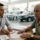 Dois homens em mesa de concessionária. Vendedor aponta documentos ao idoso com expressão surpresa.
