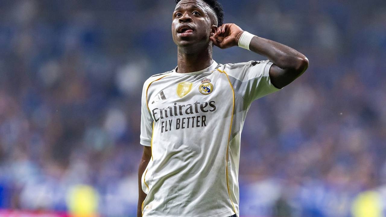 Vinicius Junior em camisa branca do Real Madrid, mão na orelha, celebrando em campo.