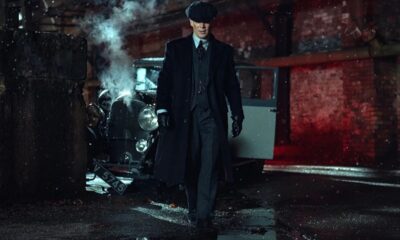 Homem de sobretudo e boina em rua molhada, carro vintage fumegante no fundo.