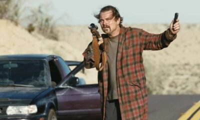 Homem com bigode e robe xadrez segura rifle e celular ao lado de carro em deserto.