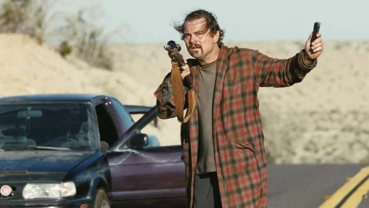 Homem com bigode e robe xadrez segura rifle e celular ao lado de carro em deserto.