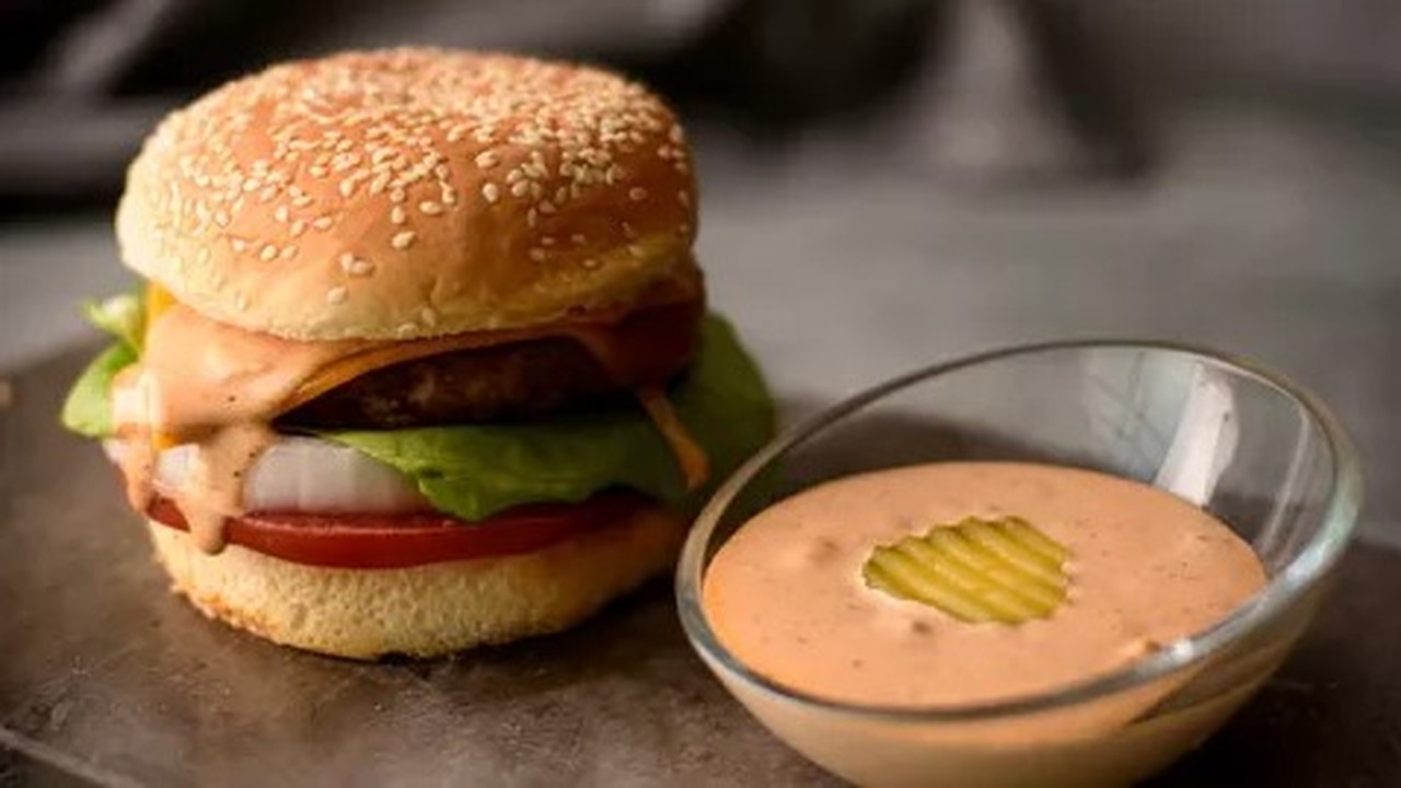 Hambúrguer completo com queijo e molho, ao lado de tigela com molho laranja e picles.