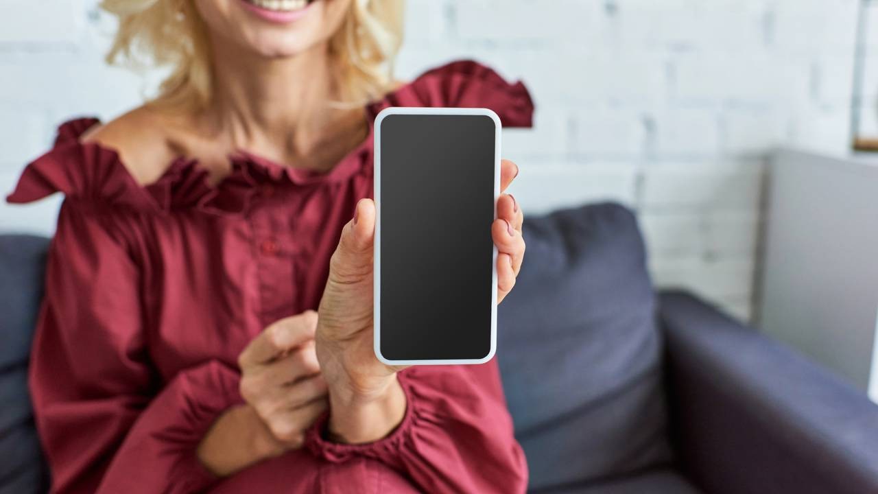 Pessoa sorridente segura smartphone branco com tela preta vazia, mostrando-o à câmera.