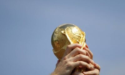 Taça dourada da Copa do Mundo sendo segurada por mãos contra um céu azul claro.