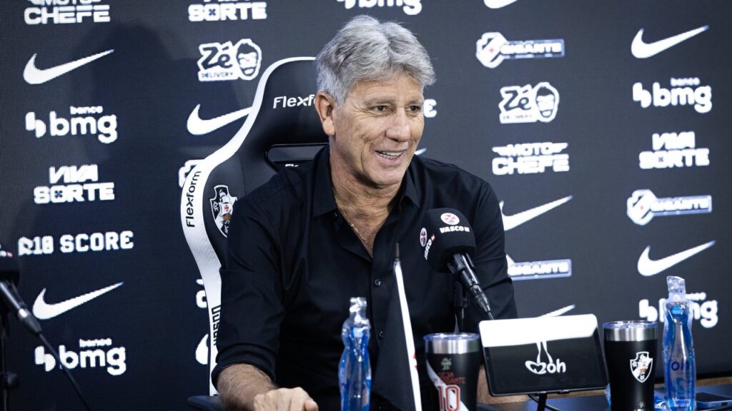 Renato Gaúcho sorri em coletiva, microfone Vasco, logos patrocinadores, garrafa e copos na mesa.