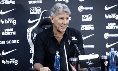 Renato Gaúcho sorri em coletiva, microfone Vasco, logos patrocinadores, garrafa e copos na mesa.