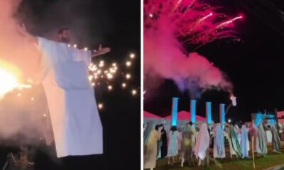 Ator em túnica branca com fogo no figurino e cena noturna de espetáculo com fogos de artifício.