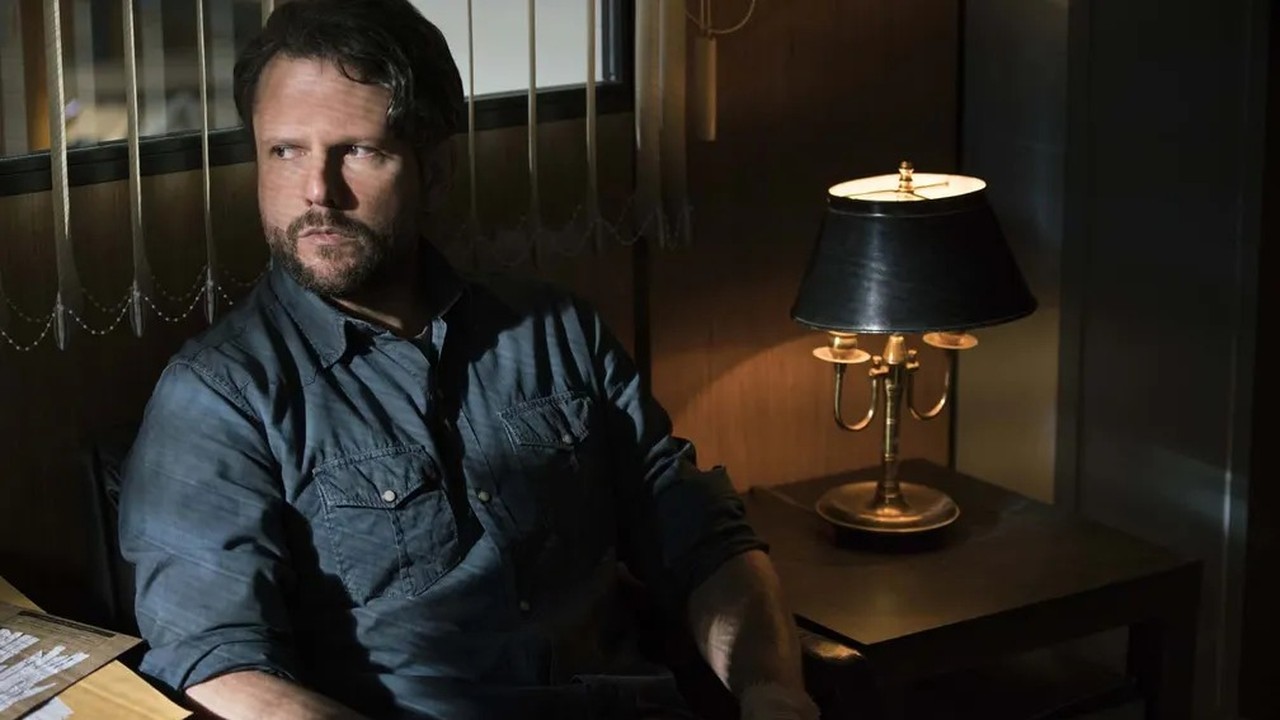 Homem barbudo de camisa jeans, olhando para a esquerda, em ambiente escuro com luminária acesa.