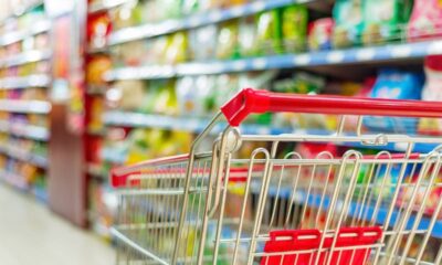 Carrinho de compras vazio em corredor de supermercado com prateleiras de produtos desfocadas.