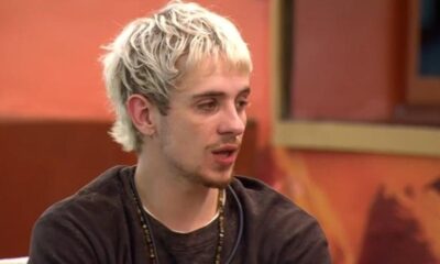 Juliano Floss, homem jovem com cabelo loiro, usa camisa escura e colar, fala com boca aberta.