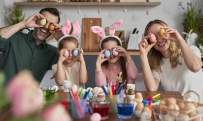 Família sorridente com ovos de Páscoa coloridos cobrindo os olhos, vestindo orelhas de coelho.