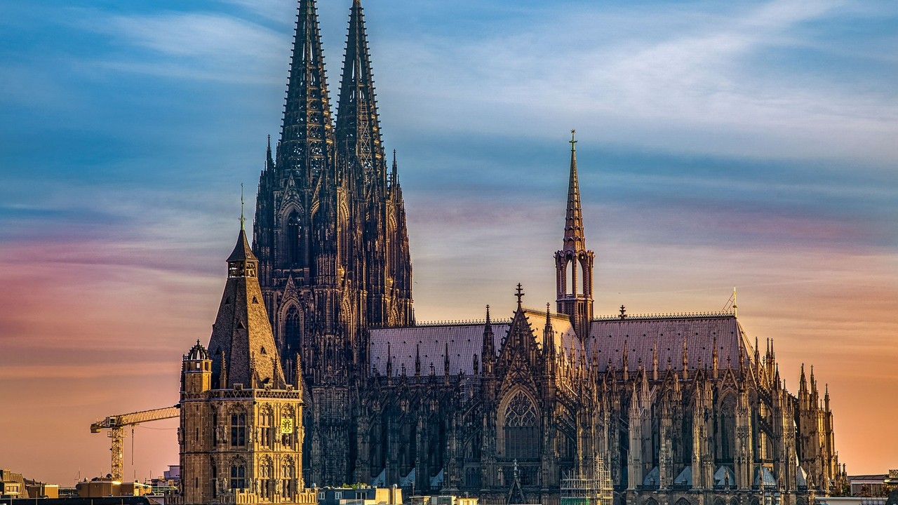 Catedral de Colônia e edifícios históricos sob céu degradê azul e laranja.