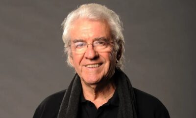 Homem idoso, Juca de Oliveira, cabelos brancos e óculos, sorrindo em fundo cinza.