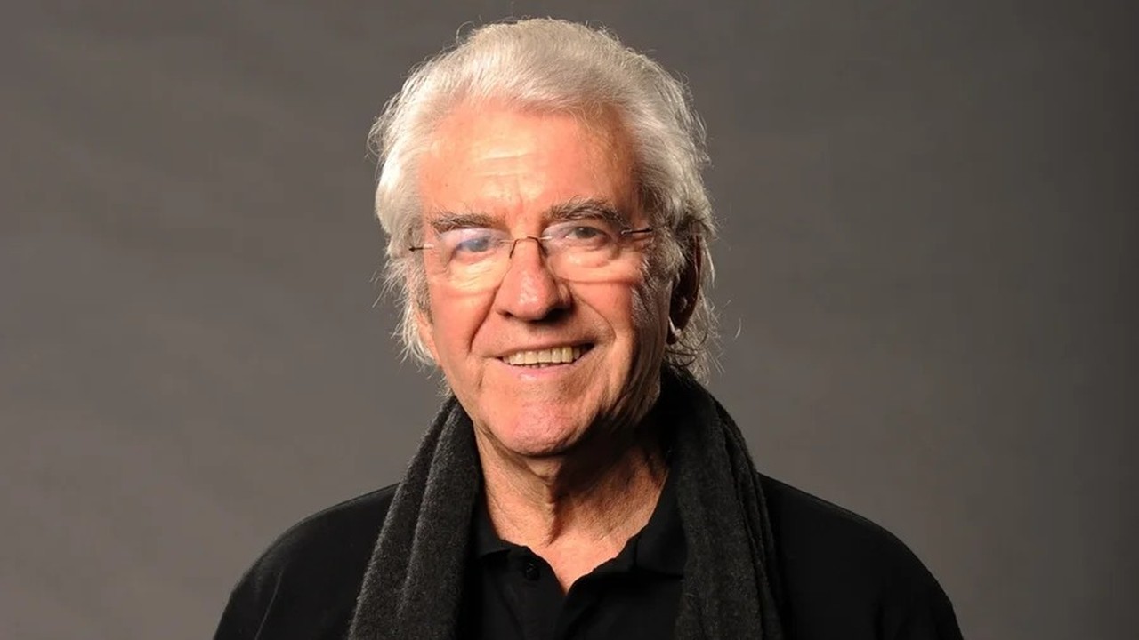 Homem idoso, Juca de Oliveira, cabelos brancos e óculos, sorrindo em fundo cinza.