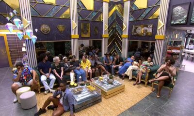 Grupo de confinados em sala de estar luxuosa com tema dinheiro, sentados em sofás e chão.