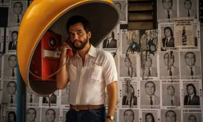 Homem barbudo fala ao telefone público vermelho, com cabine amarela e cartazes políticos no fundo.