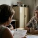 Três pessoas em casa; idosa triste à mesa com documentos de cartório.