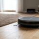 Aspirador robô cinza em piso de madeira claro ao lado de tapete marrom.