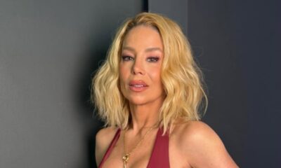 Close-up de Danielle Winits, loira, sorrindo, maquiagem rosa, blusa vinho, colar dourado.