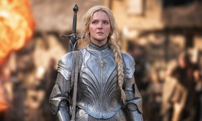 Mulher loira em armadura élfica prateada com espada nas costas, olhar determinado, fundo desfocado com fogo.