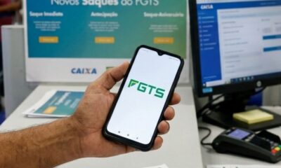 Mão segura celular com aplicativo FGTS. Ao fundo, monitor de computador e banner da Caixa.