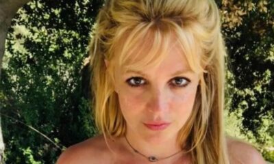 Rosto de Britney Spears, cabelo loiro com franja, colar, fundo de folhagem verde.