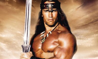 Arnold Schwarzenegger como Conan, musculoso, segurando espada, com colar e bandana, céu nublado de fundo.