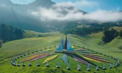 Vista aérea de arena circular com flores coloridas, estátuas, e montanhas nebulosas ao fundo.