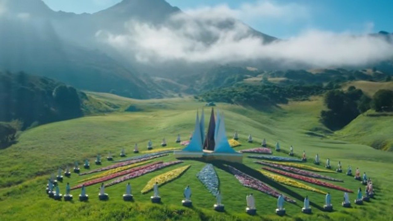 Vista aérea de arena circular com flores coloridas, estátuas, e montanhas nebulosas ao fundo.