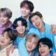 Sete membros do BTS sorriem, posando juntos em roupas coloridas vibrantes contra fundo branco.