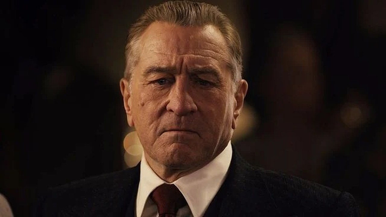Close-up de Robert De Niro, homem mais velho, rosto sério, terno escuro, camisa branca.