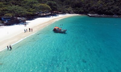 Vista aérea de praia de areia branca com mar azul-turquesa, barco e pessoas na água.