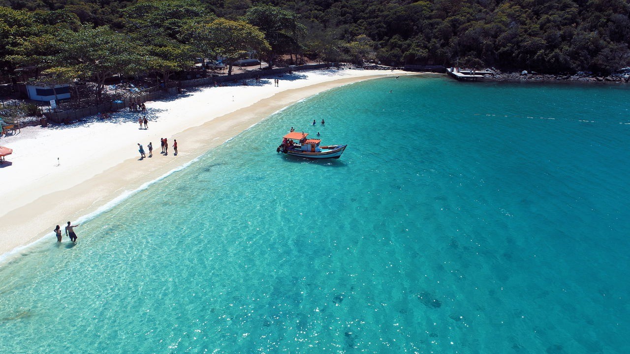 Vista aérea de praia de areia branca com mar azul-turquesa, barco e pessoas na água.